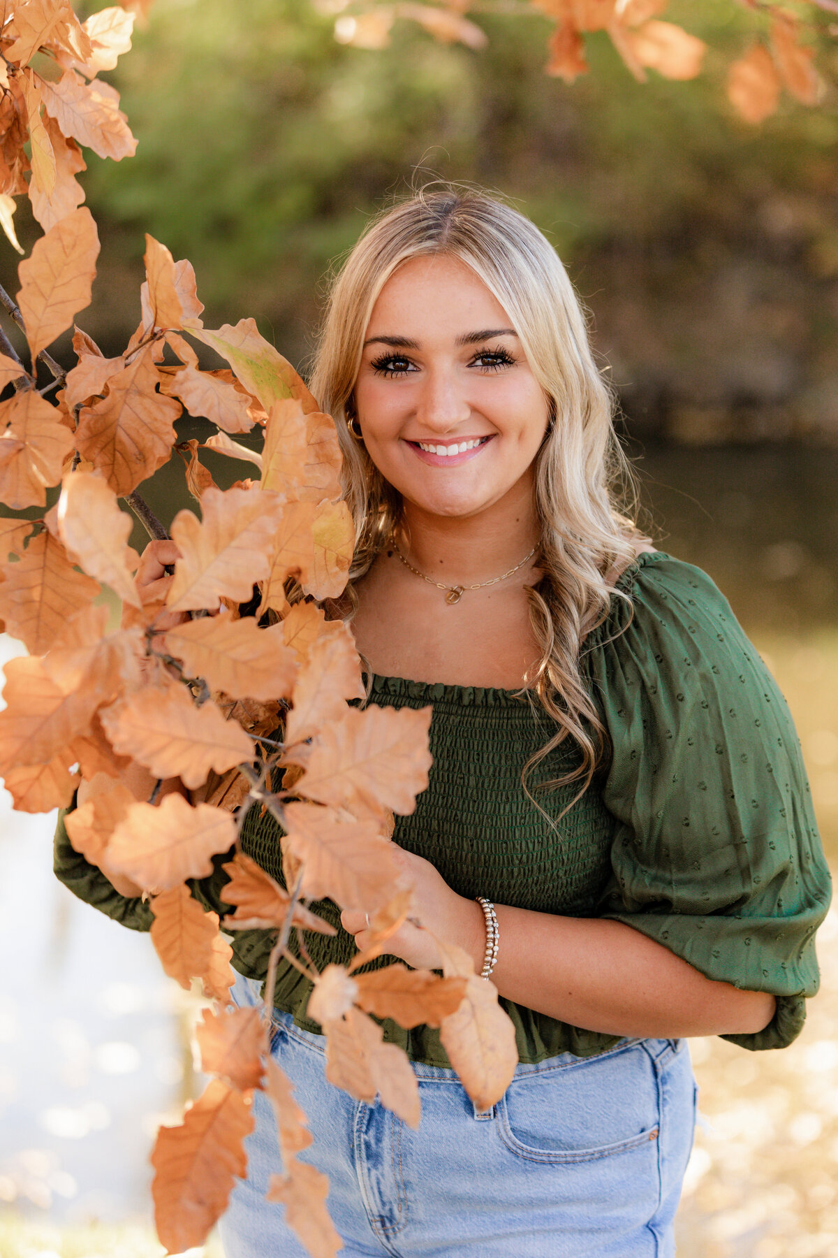 A blonde girl in a green shirt holds a branch of orange leaves in front of her for her fall senior session at lincoln landing.