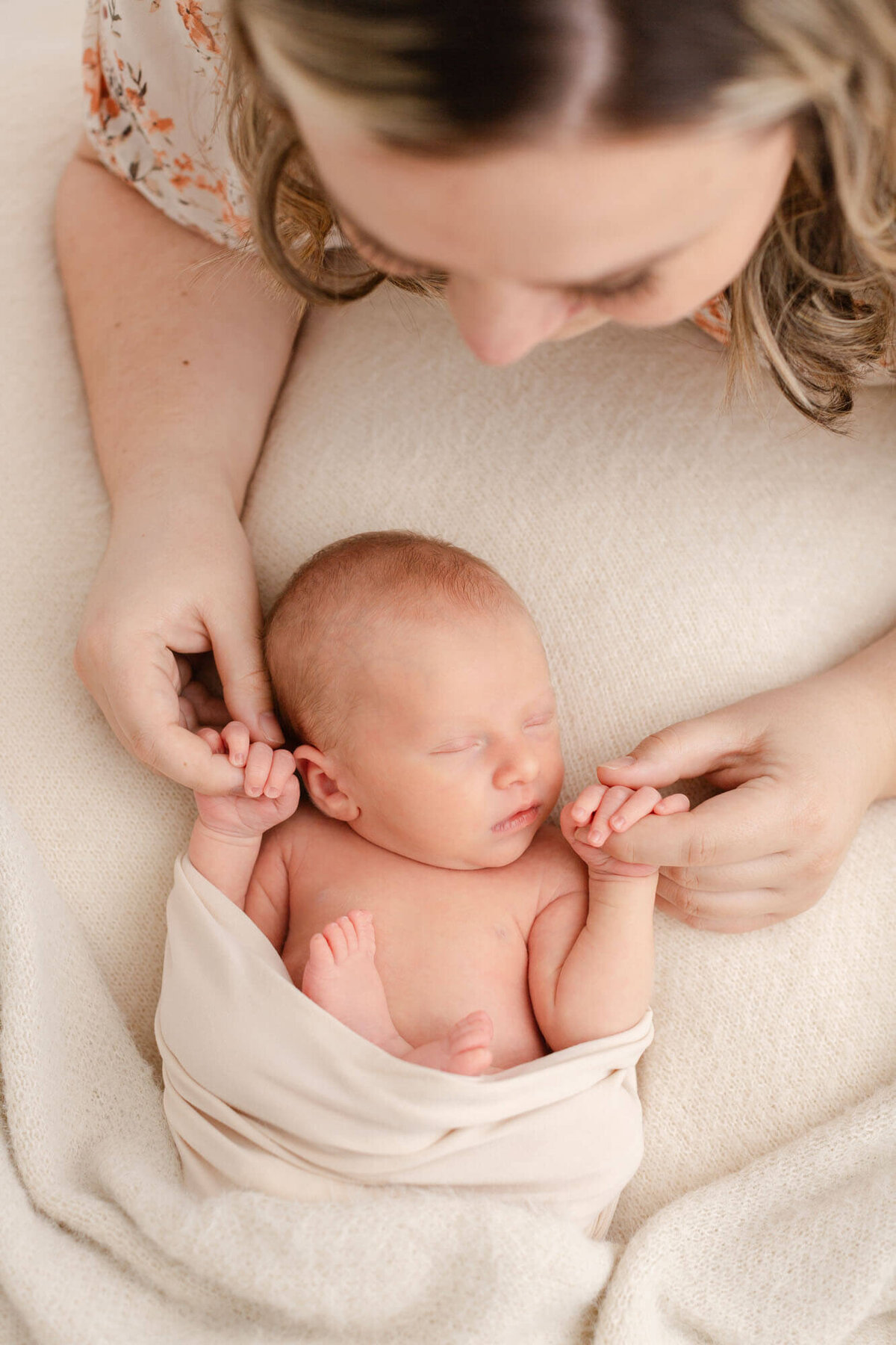 Baby is sleeping peacefully on back with arms up by face and holding Mama's hands. The top of Mama's head is visible in this image, but the focus is on the baby and Mom holding Baby's hands.