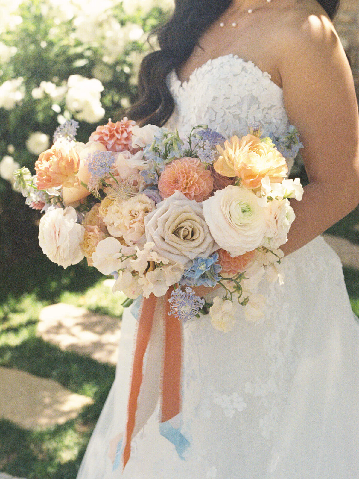 Bride in a lacy white dress holding a vibrant bouquet of roses and assorted flowers in pastels and orange tones, evoking a joyful, romantic ambiance.