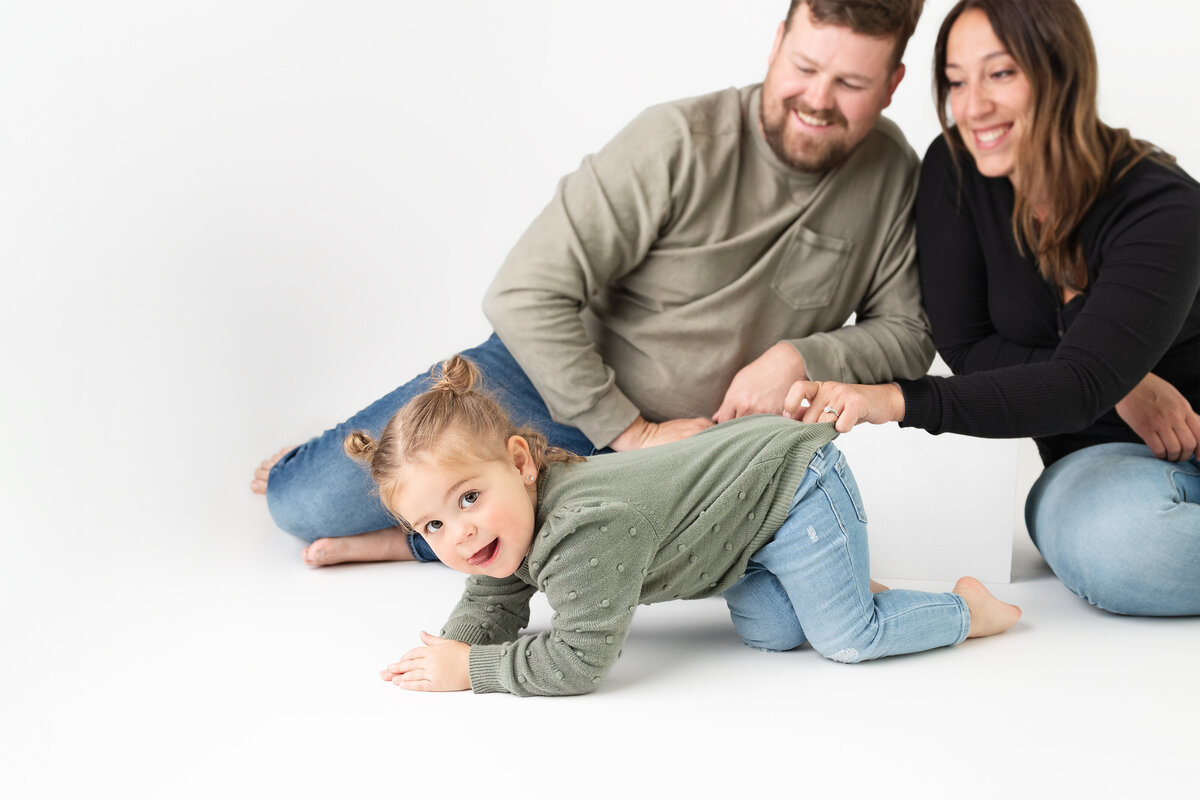 Toddler crawling and smiling toward the camera while parents laugh behind her in a white studio space in Paris, Ontario