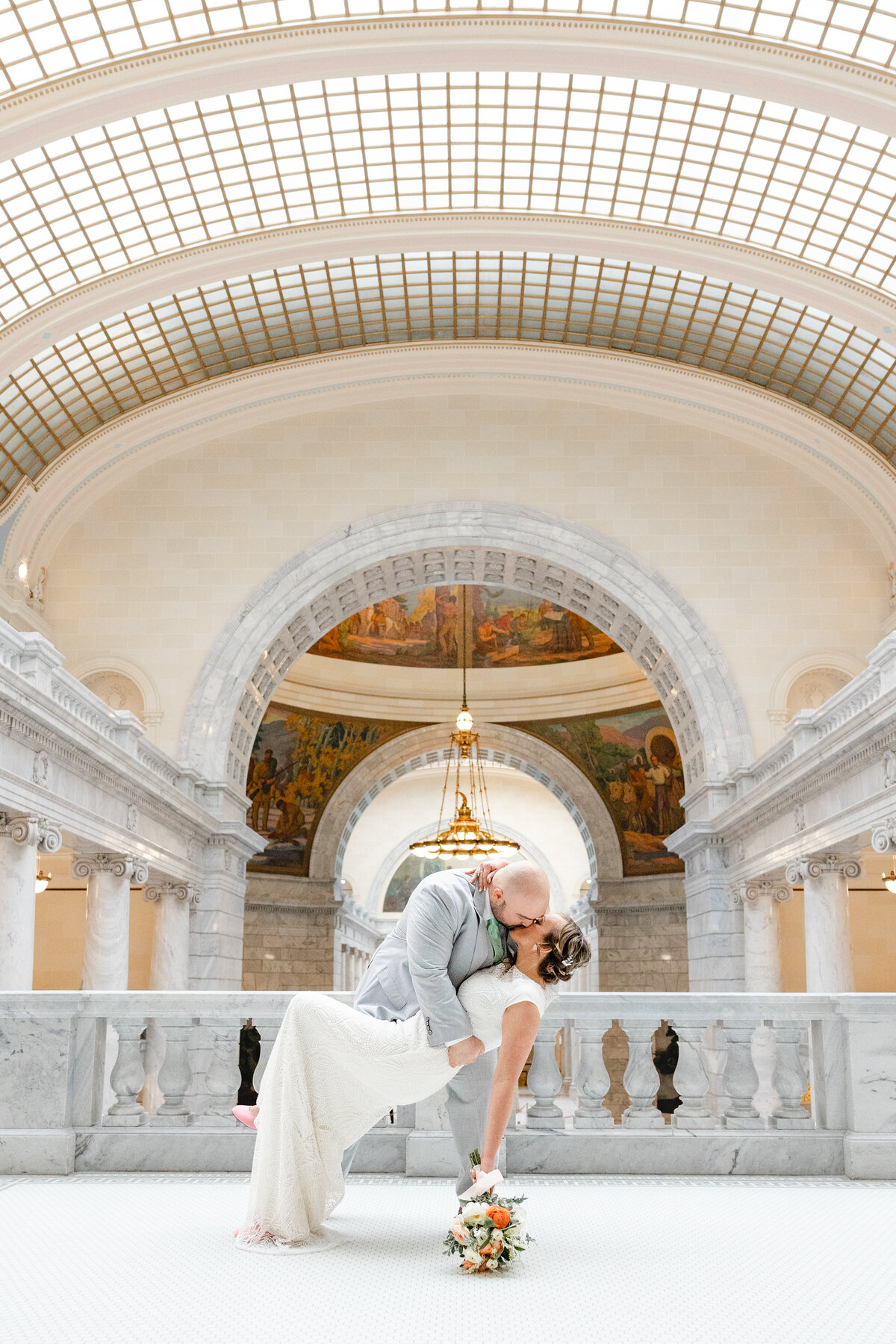 Groom romantically dipping his bride for a kiss in the elegant halls of the Utah State Capitol, with chandeliers and grand architecture in the background.