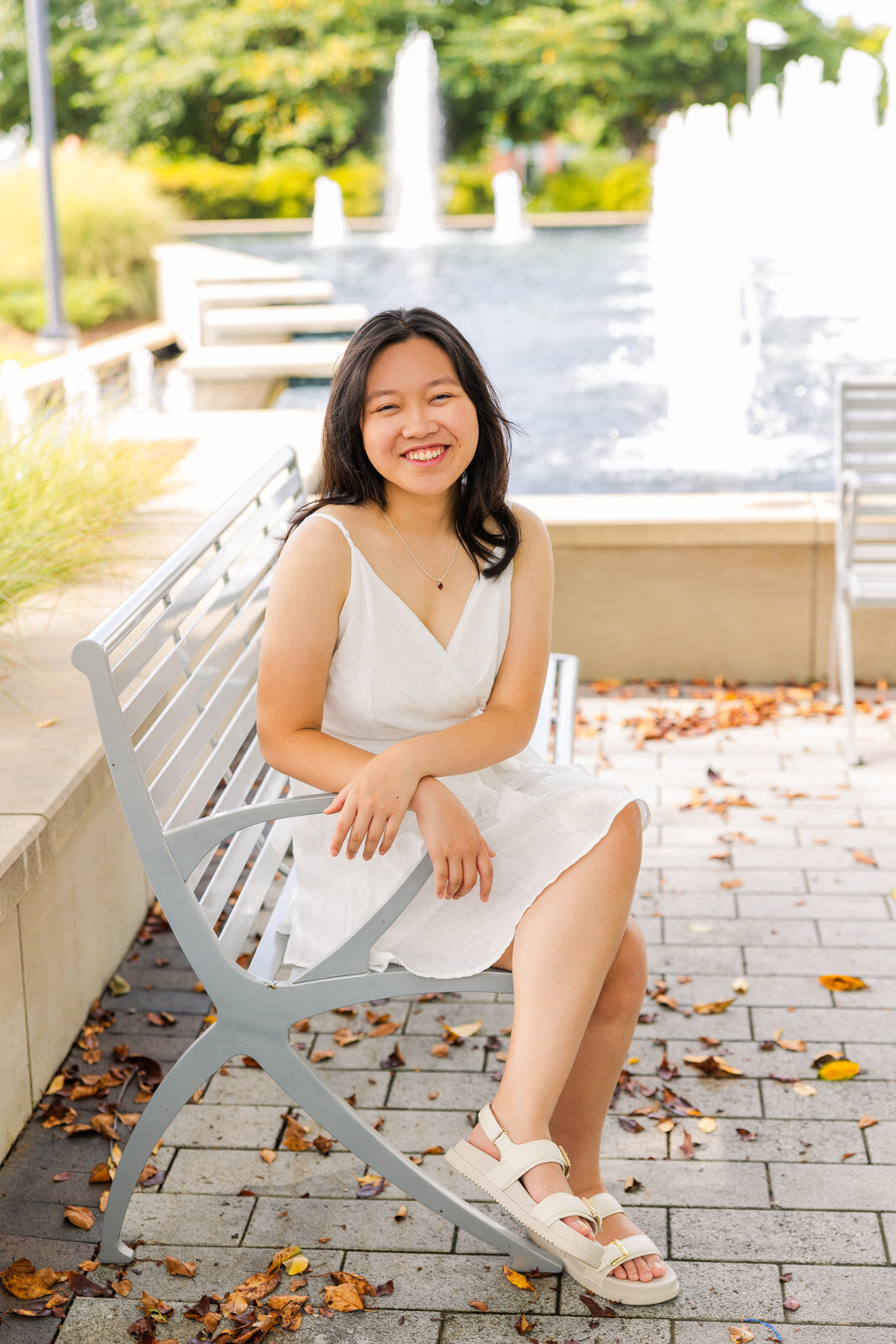 high school girl siting on a white bench in front of a fountain legs crossed and arms resting on the side during photoshoot