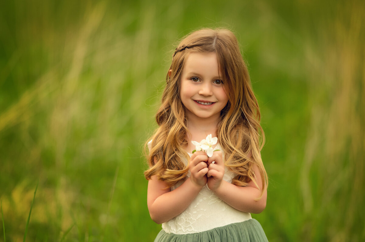 A young girl stands in a tall field at Minooka Park holding a small white flower while her portrait is taken.