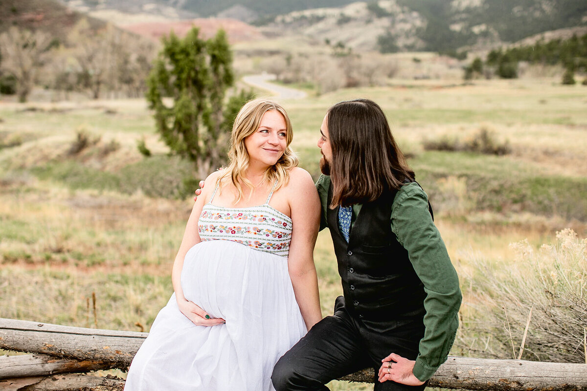 pregnant woman in white dress and her partner look at each other while sitting on a fence