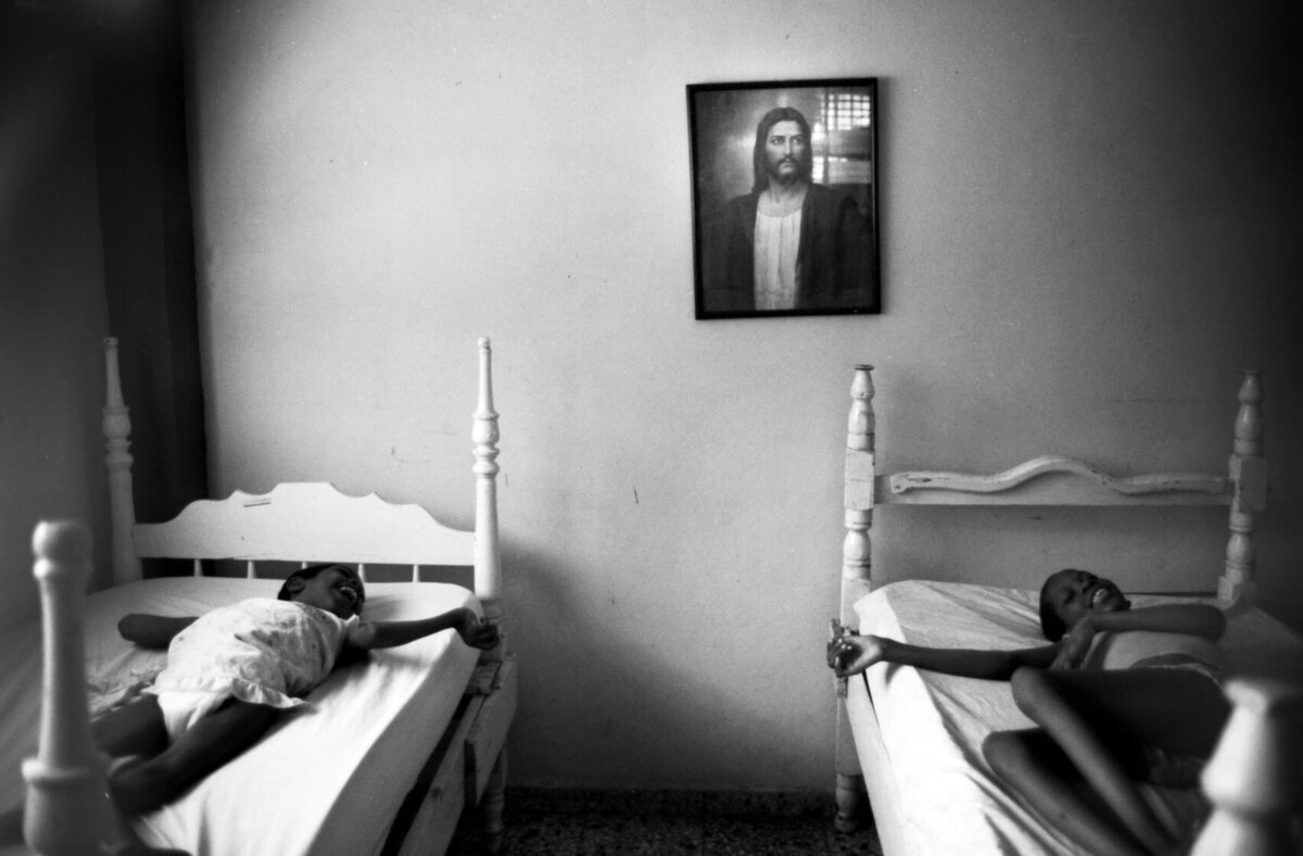 Children reach for each-other with image of Jesus on wall above them.  Shot in black and white