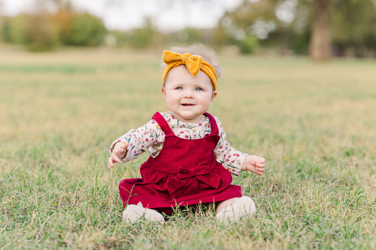 baby-girl-wearing-a-red-dress-with-gold-bow-in-her-hair-sitting-in-a-green-grass-filed