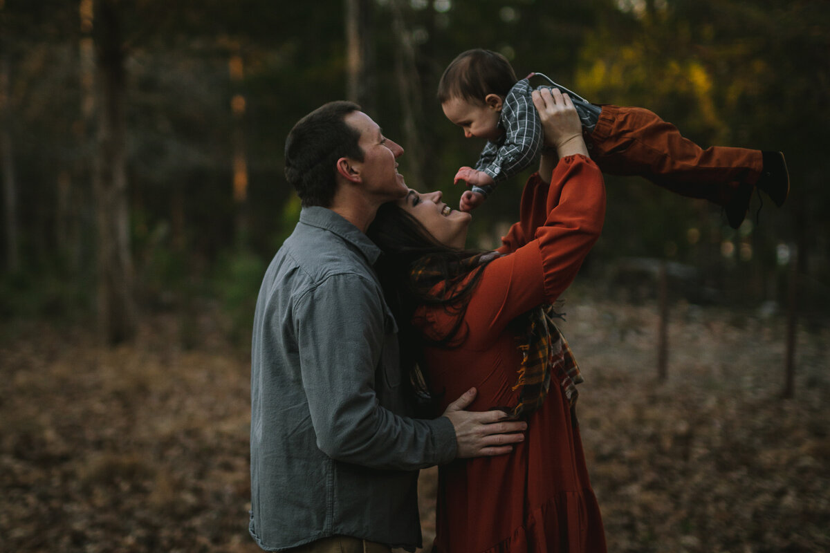 Father holds mother by the waist as she lifts their son over her head and they both smile as he smiles back at them.