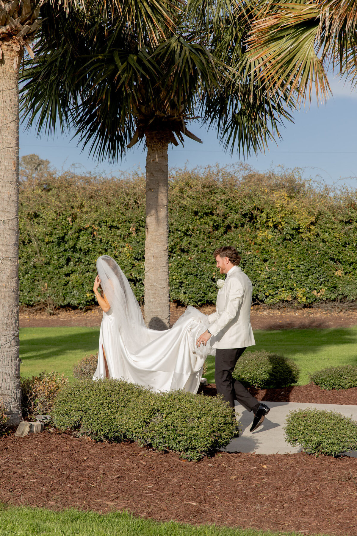 A groom carries the train for his bride during their wedding ceremony. They're outside with lots of greenery and large hedges behind them.
