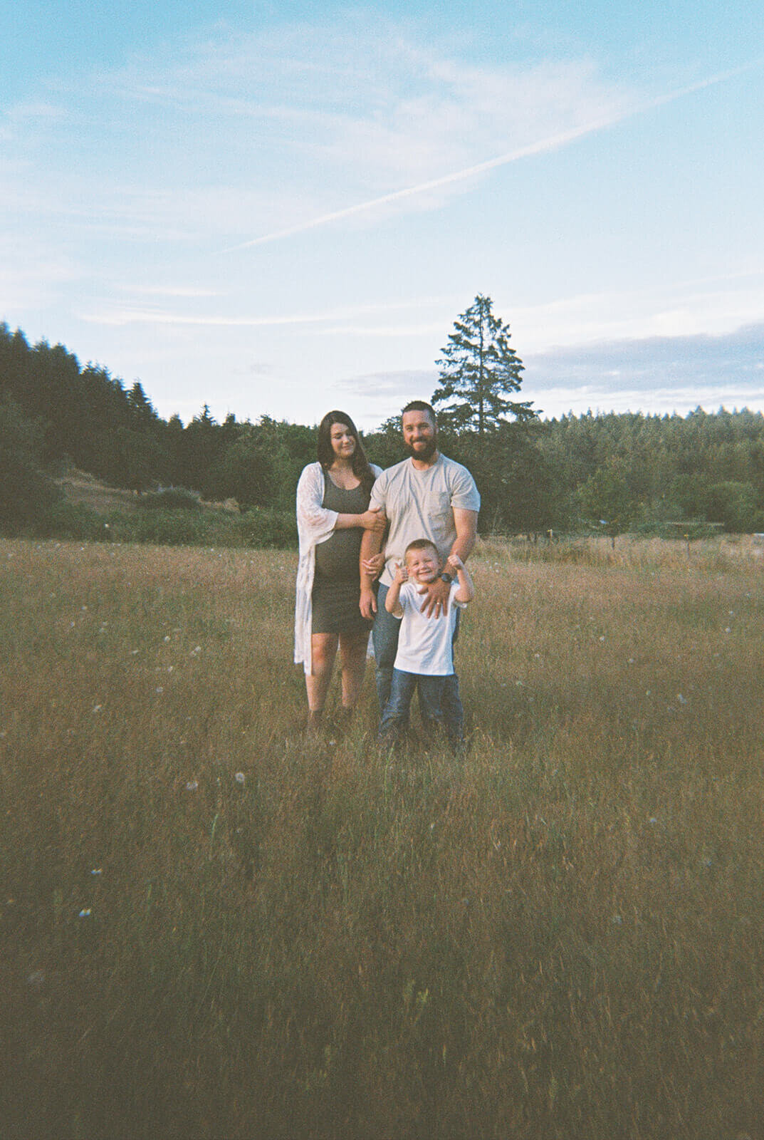 A family of three stands in a field during a maternity photography shoot.
