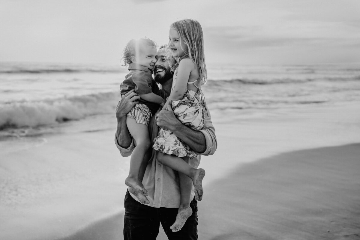 Father smiling while holding his two young children in his arms on the beach, black and white San Diego family photo.