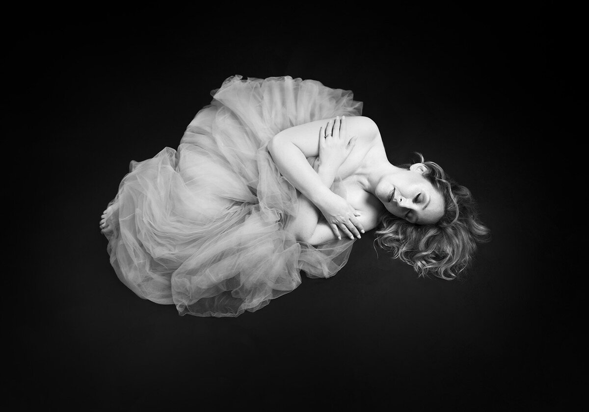 A woman in a tulle shirt laying curled up on the floor with ther hair flowing. Her arms are crossed across her chest. Taken by Norwich boudoir photographer Claire Howes.
