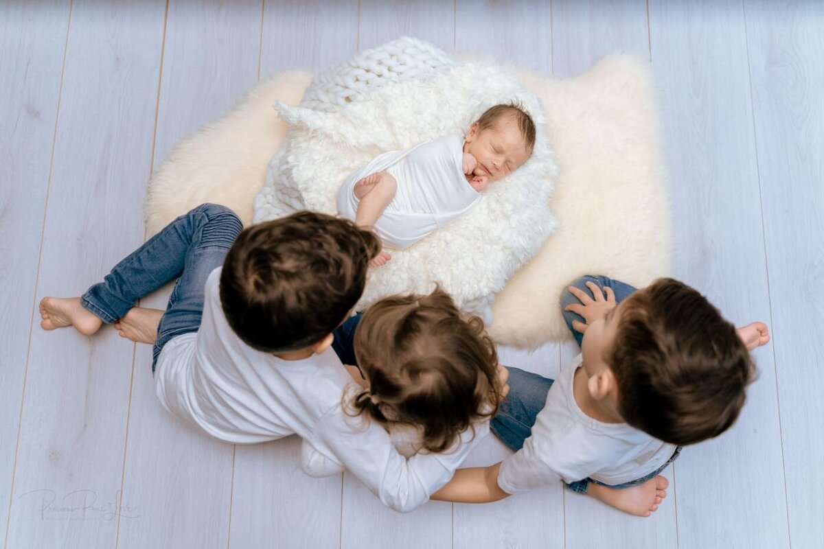 Siblings with newborn three young siblings sitting barefoot around their sleeping newborn brother, who is wrapped in white and lying on a fluffy cream blanket on light wooden floor.