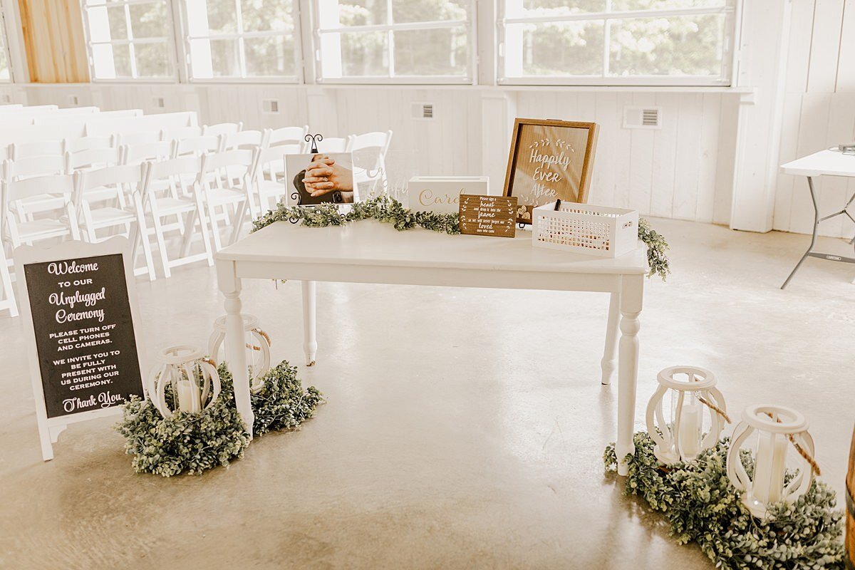 Inside a white ceremony barn at Grace Valley Farm sits a white farm table topped with light brown signs welcoming guests and asking them to sign the guests book. On the floor at either side of the table are white lanterns wreathed in greenery and an unplugged wedding sign sitting to the left of the table.