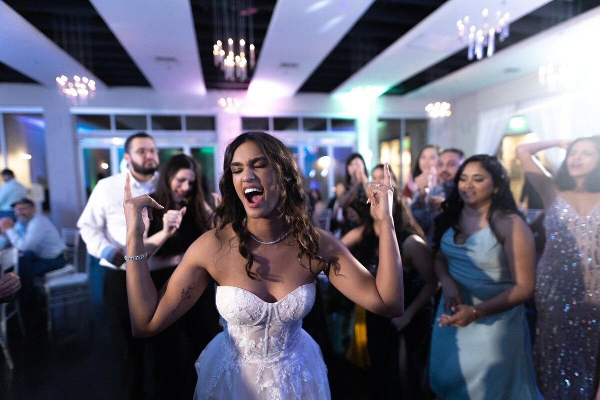 A bride in a sleek white gown stands beside her seated groom in a modern lounge space at a Chandler wedding and event venue. The couple poses in front of a stylish wood bookshelf with curated décor, combining elegance with a contemporary indoor setting.