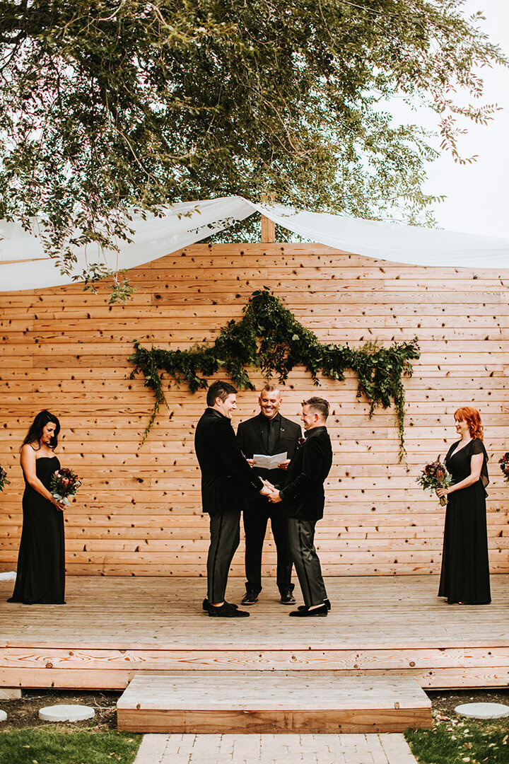 Two grooms wearing black tuxedos share a smile during their wedding in front of the officiator on a wooden stage.