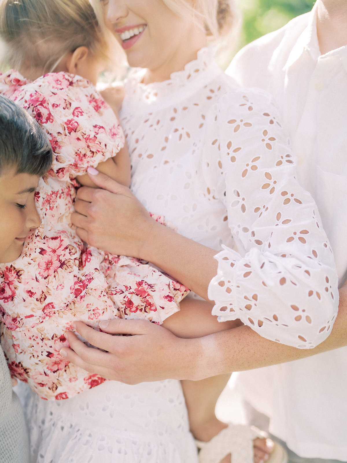 Close-up view of mother holding her toddler daughter in pink floral dress as her sons lean into her.