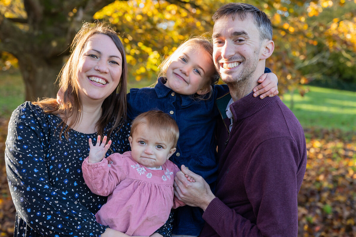 A family of four smiling in a park during autumn, with two adults and two young children, one being held and the other waving at the camera.