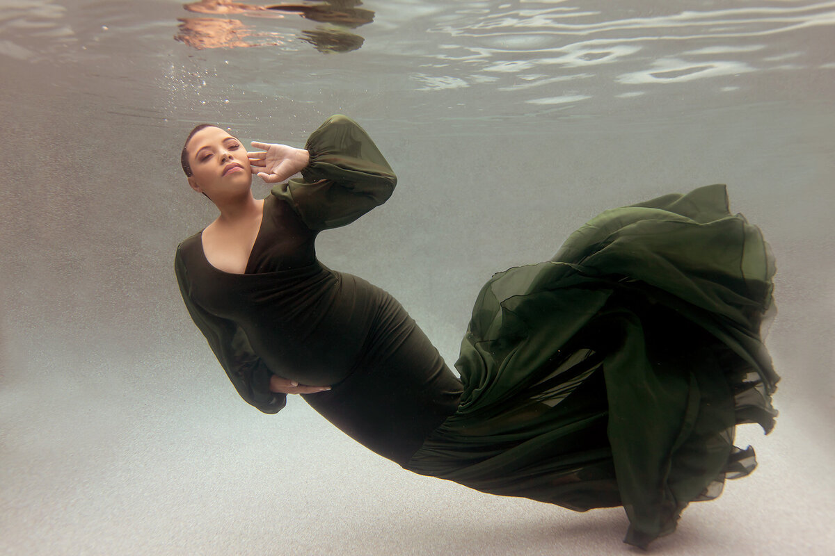 Underwater maternity photo featuring a woman in a flowing dark green gown gracefully floating in the water. Captured by a Dallas underwater pregnancy photographer, the serene setting highlights the elegant fabric swirling around her, creating a timeless, ethereal feel.
