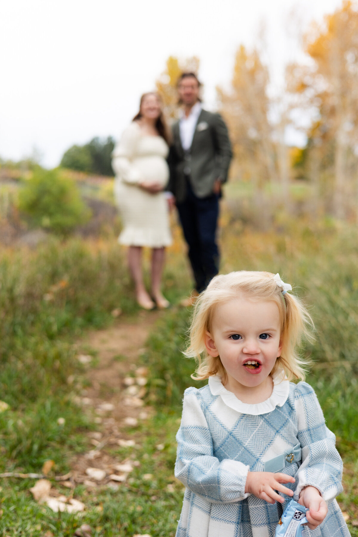Mom and dad smile at their toddler daughter in the background while she's eating a snack right in front of the camera.