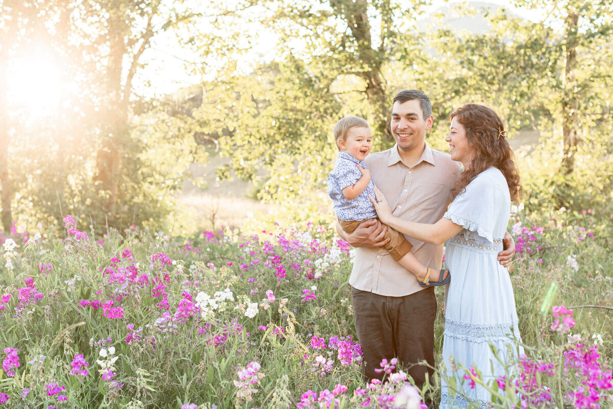Dad holding toddler son and mom standing right next to them. They are all standing in a field of pink wildflowers while the sun is glowing off to the side of them. they are all laughing together. They are wearing tones of beige, khaki and blue.