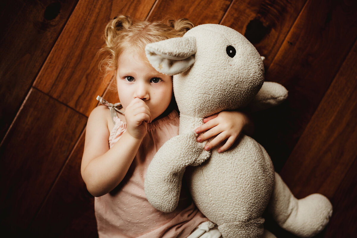 A little girl is sucking her thumb and holding her stuffed lamb while lying on the wood floor.