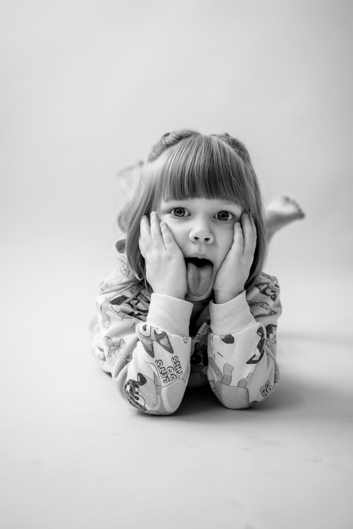 Black and white photo of girl laying on stomach with hands on her face and sticking her tongue out in studio in Severn Maryland