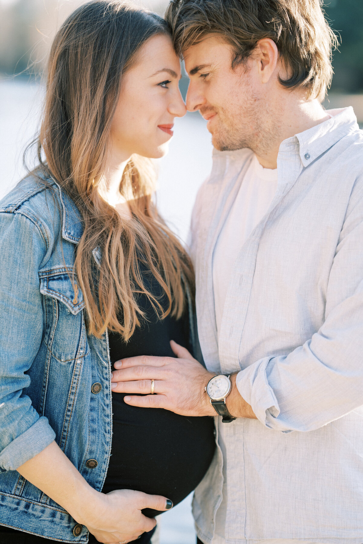Portrait of a man and a pregnant woman in a black shirt and blue jean jacket leaning into each other holding her belly.