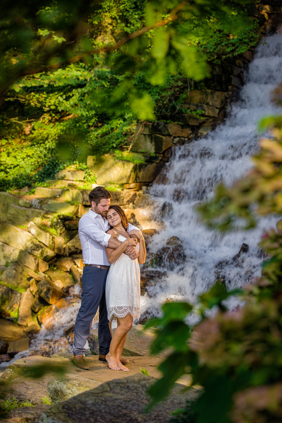 Romantic summer engagement photo of a couple standing by the cascading waterfall at Longwood Gardens, framed by lush green foliage and colorful blooming flowers.