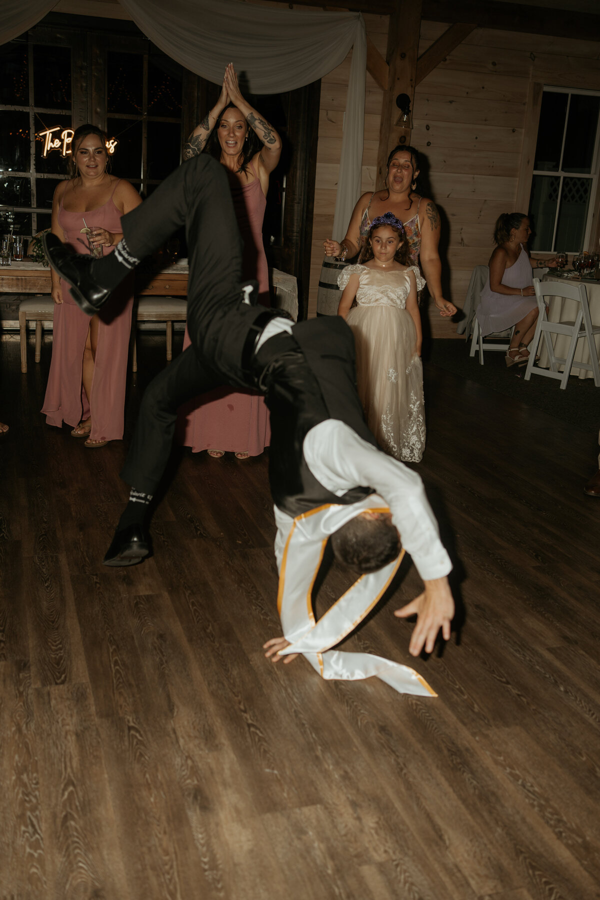 Wedding guest performs an impressive breakdance move on the dance floor, captivating the attention of other guests who cheer and applaud. The lively scene includes guests in elegant dresses and a young girl in a flower girl dress, highlighting the fun and energetic atmosphere of the celebration.