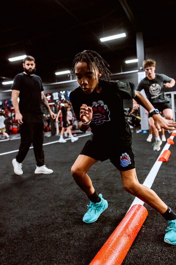 Boys football teams train on a black indoor turf, with his coach looking on.