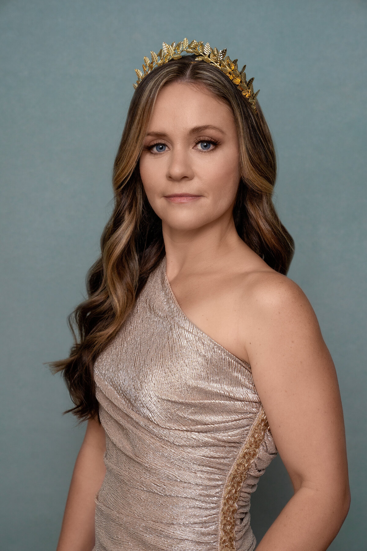 Woman wearing gold dress on blue backdrop with wavy hair and crown. 