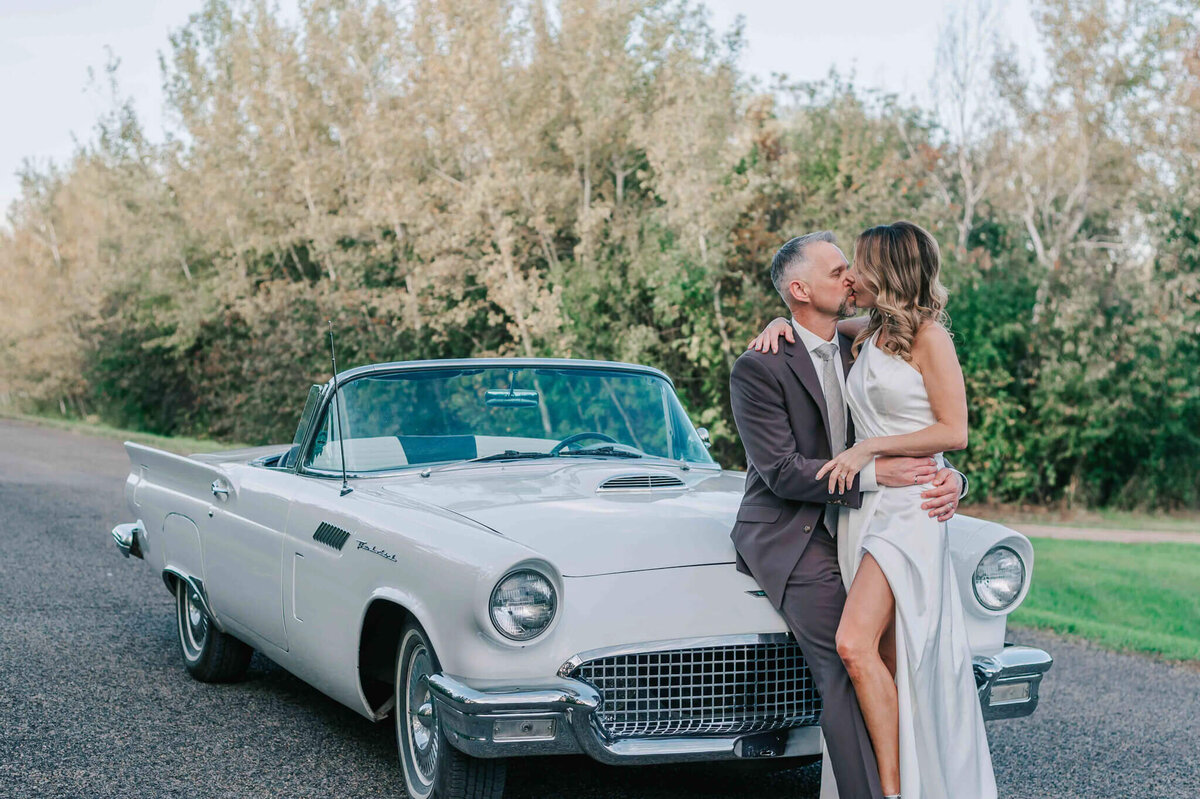 Bride and groom kissing and sitting on the hood of a white car.