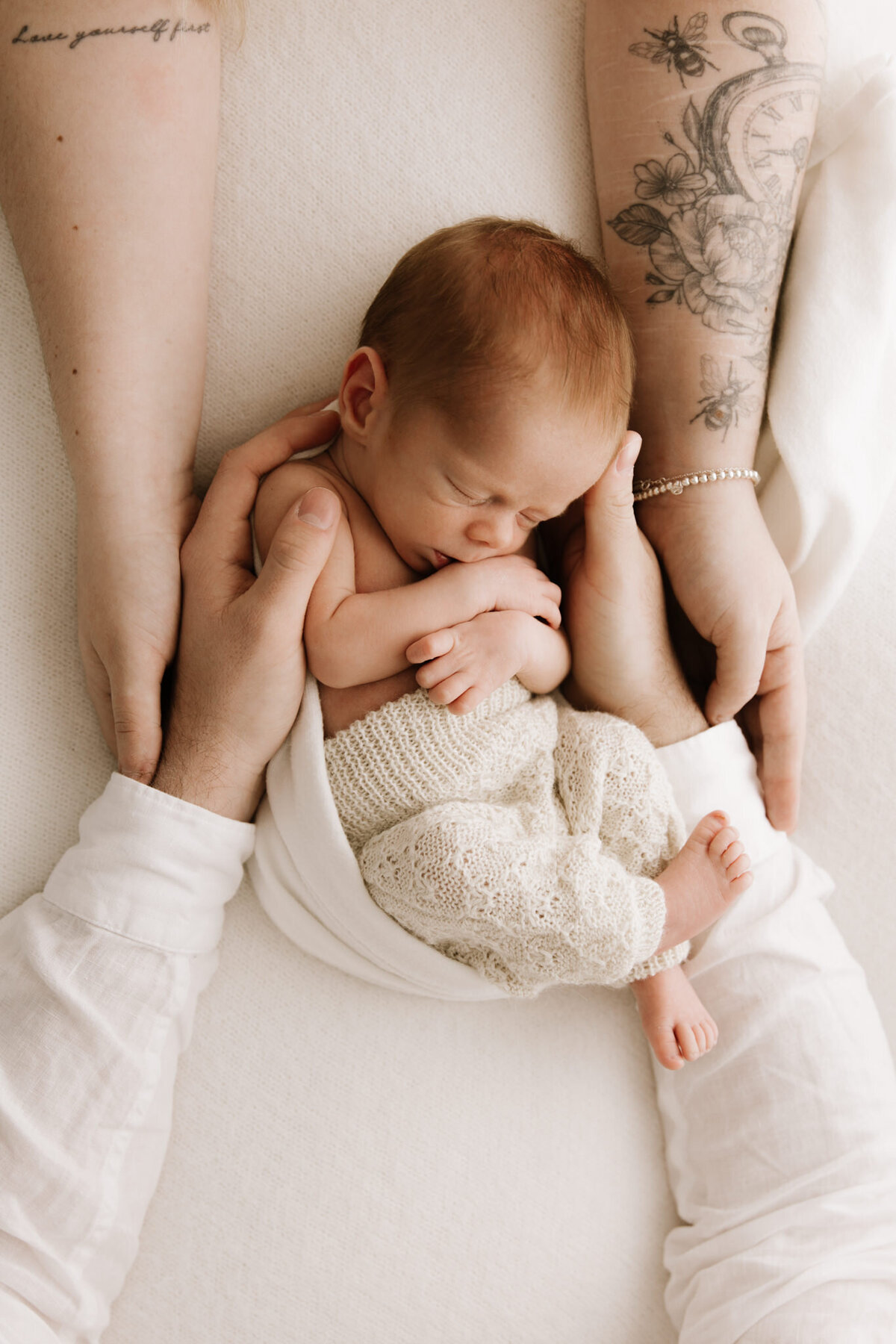 Close-up of a newborn baby wrapped in a textured cream knit outfit, peacefully asleep while cradled in the loving hands of both parents. The image highlights the intricate tattoos on the mother's forearms and a bracelet, contrasting with the softness of the baby's skin. Captured in a Sydney newborn photography studio, this image showcases warmth, unity, and intimate family connection.