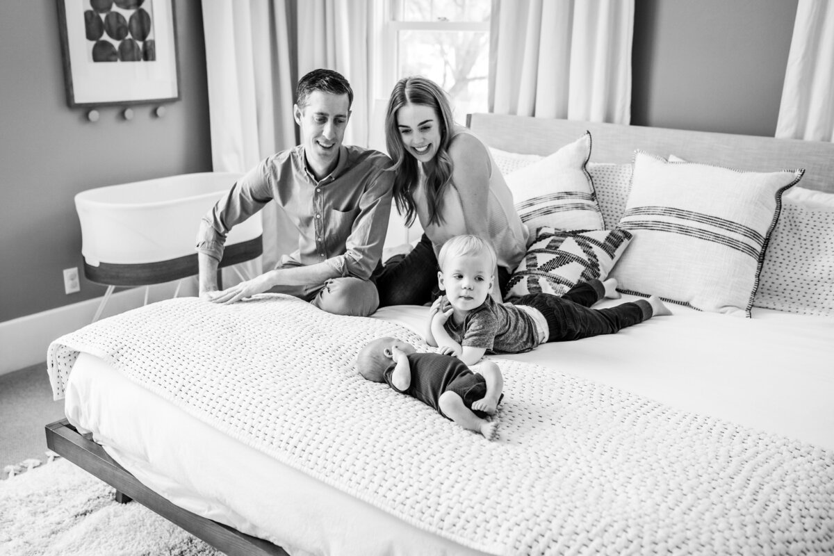 A family of four, including a toddler and newborn, relaxes on a bed with light-colored bedding and decorative pillows.