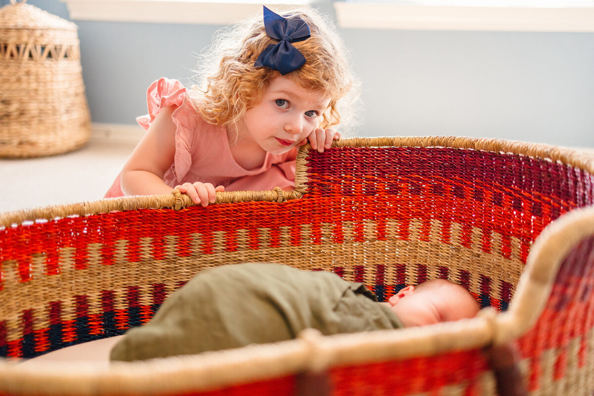 Photographs of siblings in a blue room. The little girl is a curly blonde, and is looking at the camera with a blue bow tie. The newborn baby is sleeping with a green blanket