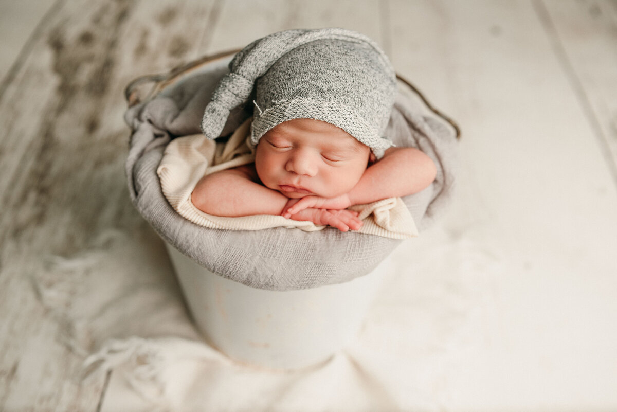 Baby boy newborn posed in white bucket with chin on hands sleeping with gray sleepy cap on