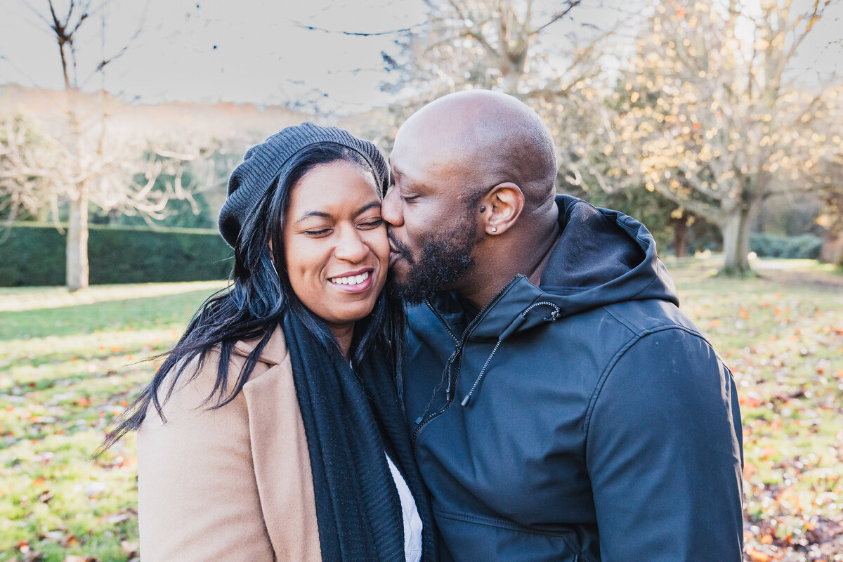 A man kissing a smiling woman on the cheek in a park, both wearing winter clothes, with sunlight and fallen leaves around.