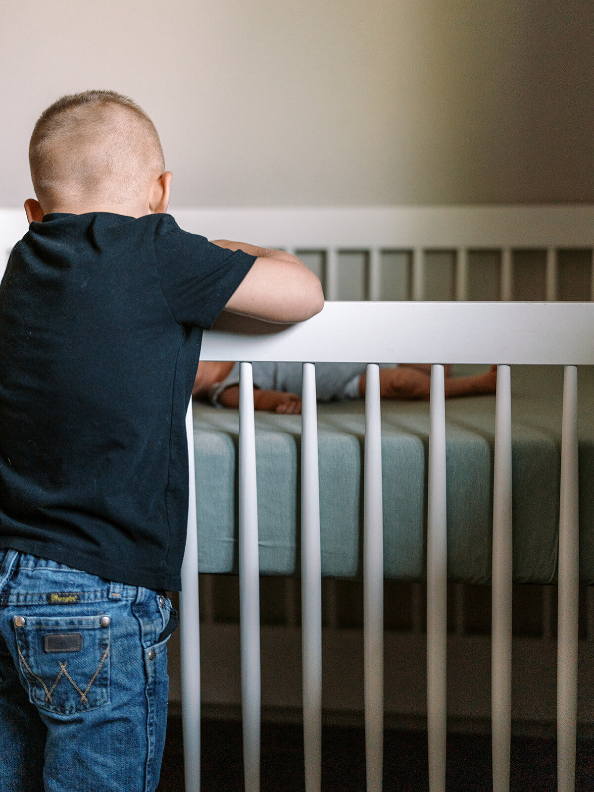 A young boy looking over the side of a crib at his newborn sibling.