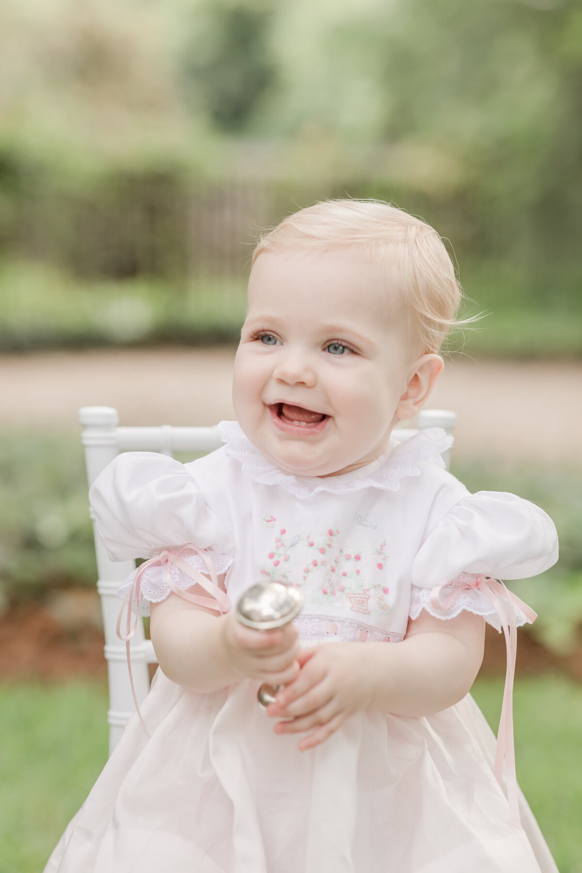 One year old girl holding a silver rattle and beaming for a portrait.