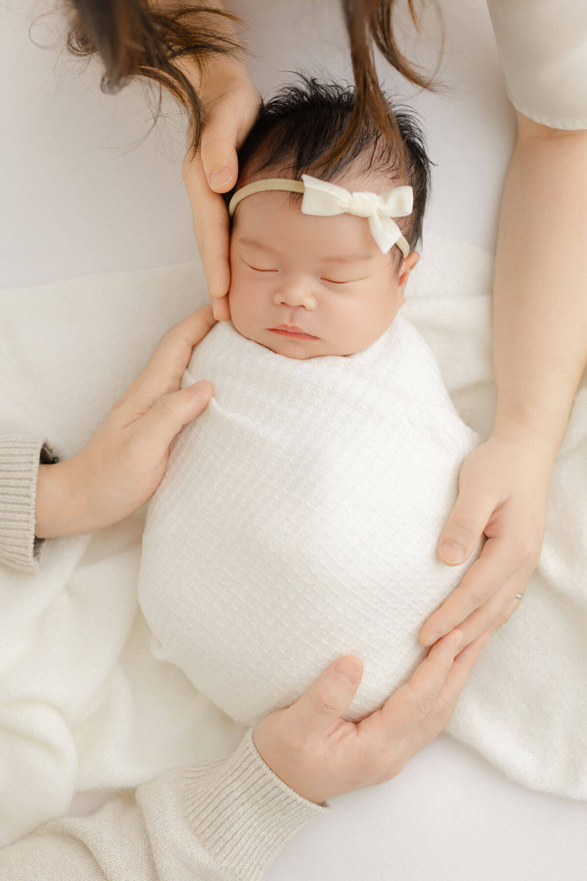 Baby girl wrapped in white with parents hands in the image holding her while she lays on a white blanket.