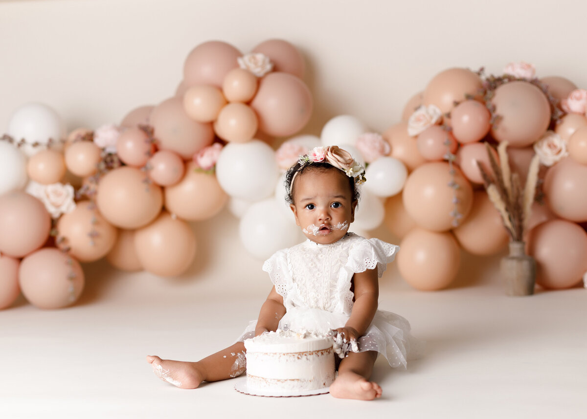 Beautiful boho desert themed cake smash at top West Palm Beach, FL and Jupiter, FL newborn and cake smash photographer. Baby girl is sitting in front of a white cake in a white lace dress, touching the cake with cake on her face. In the background there are various shades of monochromatic peach, tan, and dusty rose balloons and woven with cream flowers and feathers.