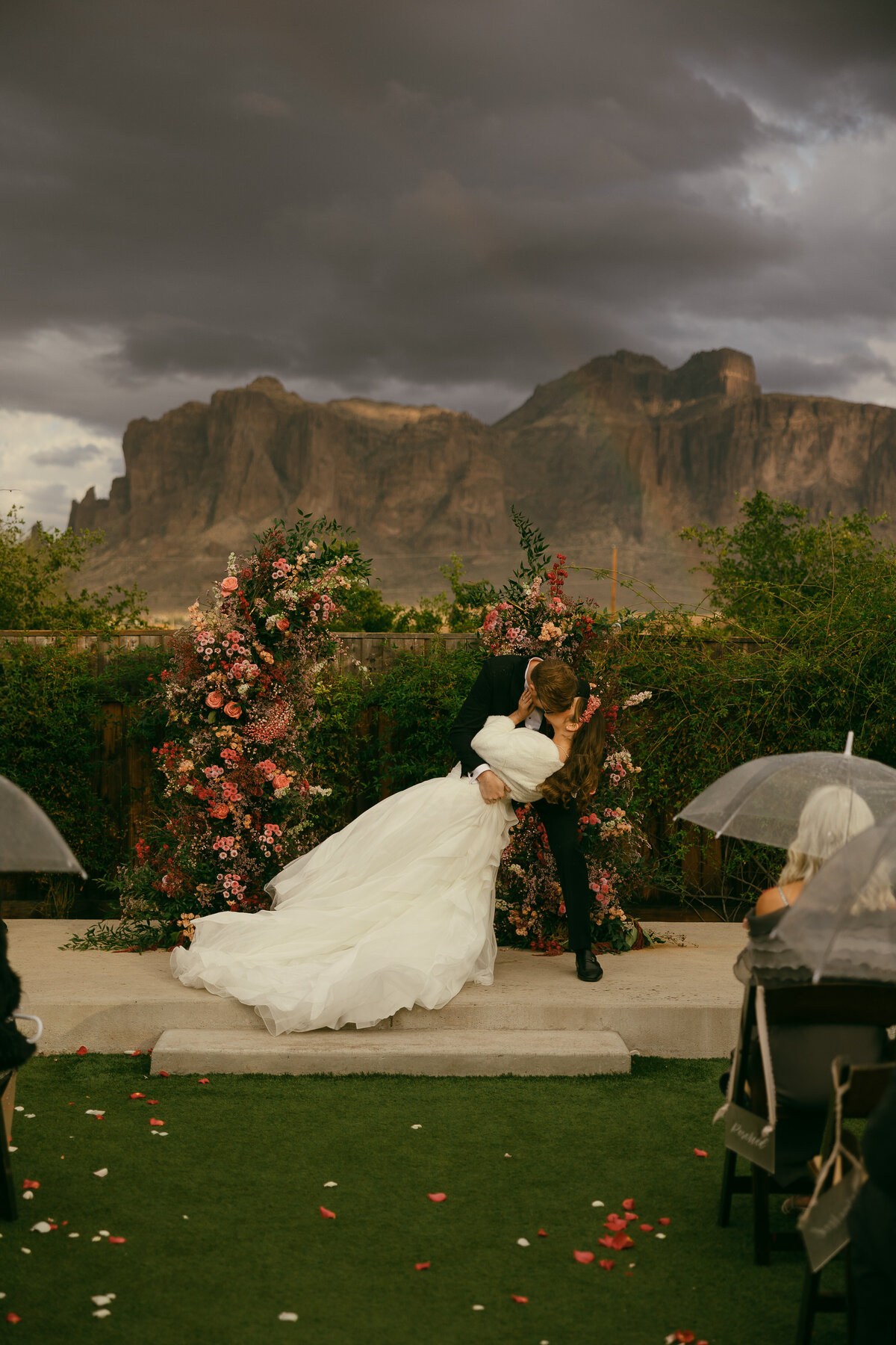 a groom dips his bride in front of an arch of multi colored florals with a stormy sky and a rainbow in front of the mountain