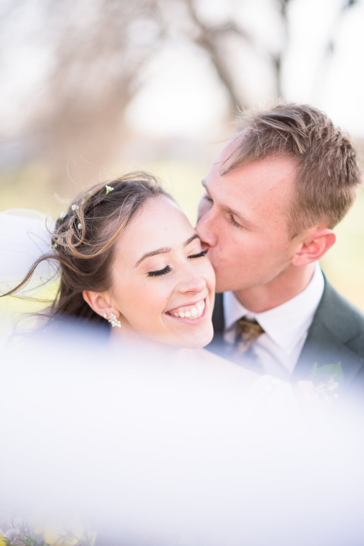 denver wedding photographer captures groom kissing his brides cheek as she smiles with her eyes closed and her veil blows in front of them