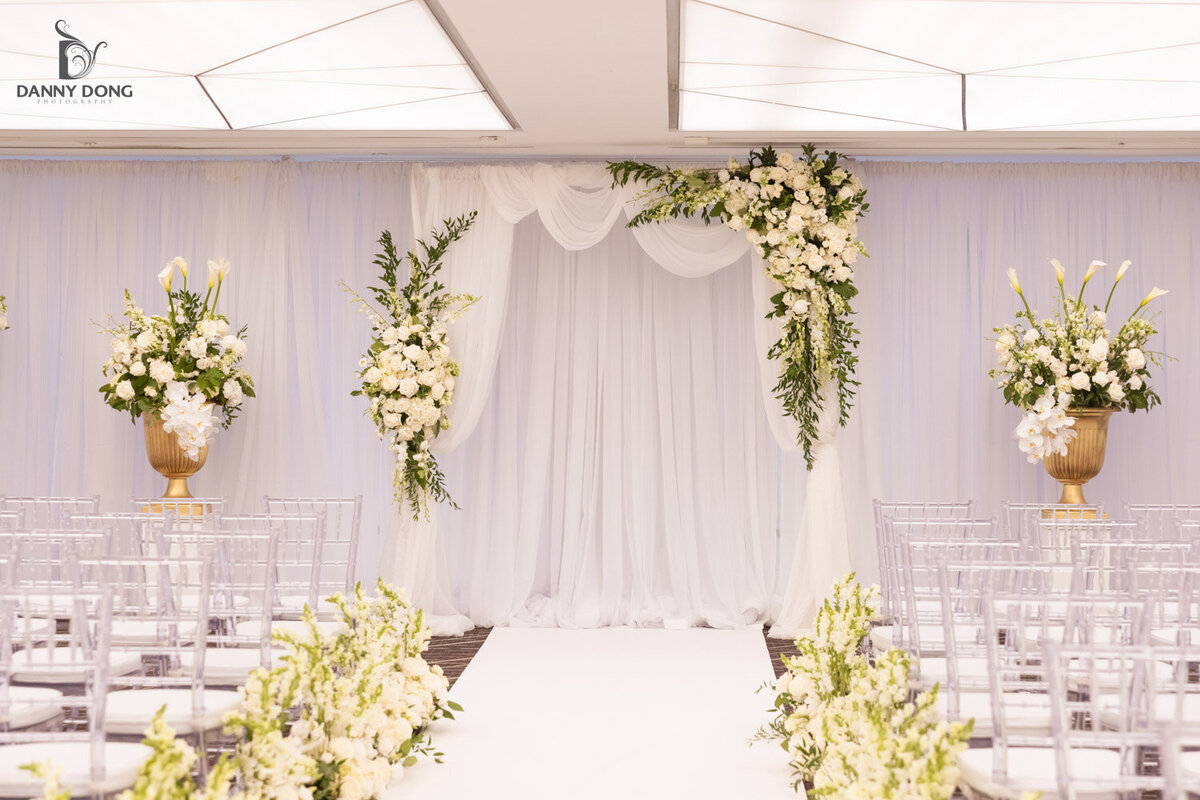 Indoor wedding ceremony aisle with white floral arch and draped backdrop