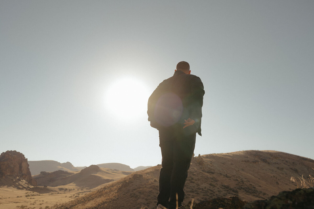 man in jacket stands on rocks as he looks out at the sunny landscape