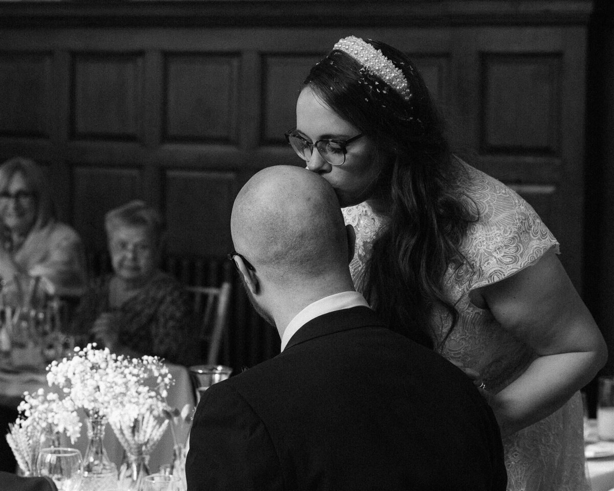 A standing bride kisses the head of her seated groom during the speeches.