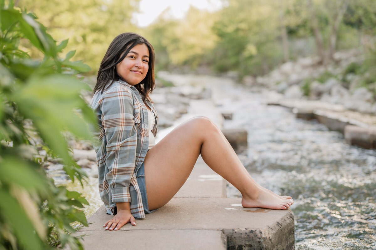 Girl sits along the edge of the river and smiles at the camera.