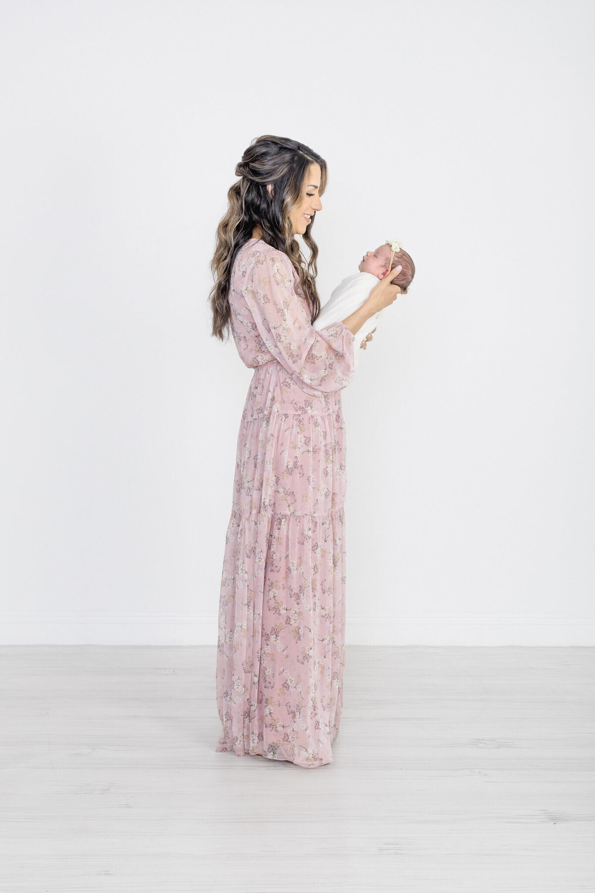 A woman in a long, pink floral dress stands in a white room, holding a newborn baby wrapped in a light blanket. She is smiling at the baby, showcasing a serene and loving moment.