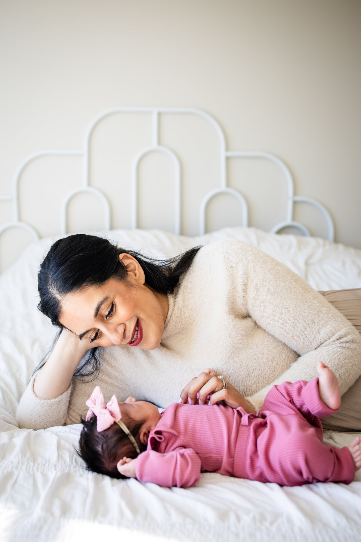 Mom laying on bed in brightly lit room with baby girl in pink outfit