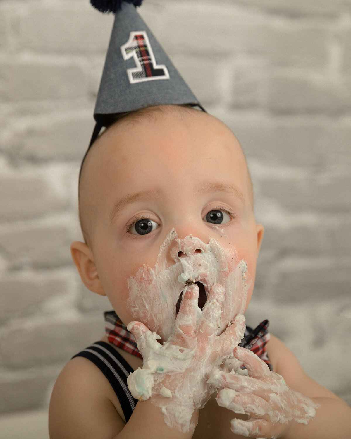 1 year old boy with blue suspenders and birthday hat eating cake.